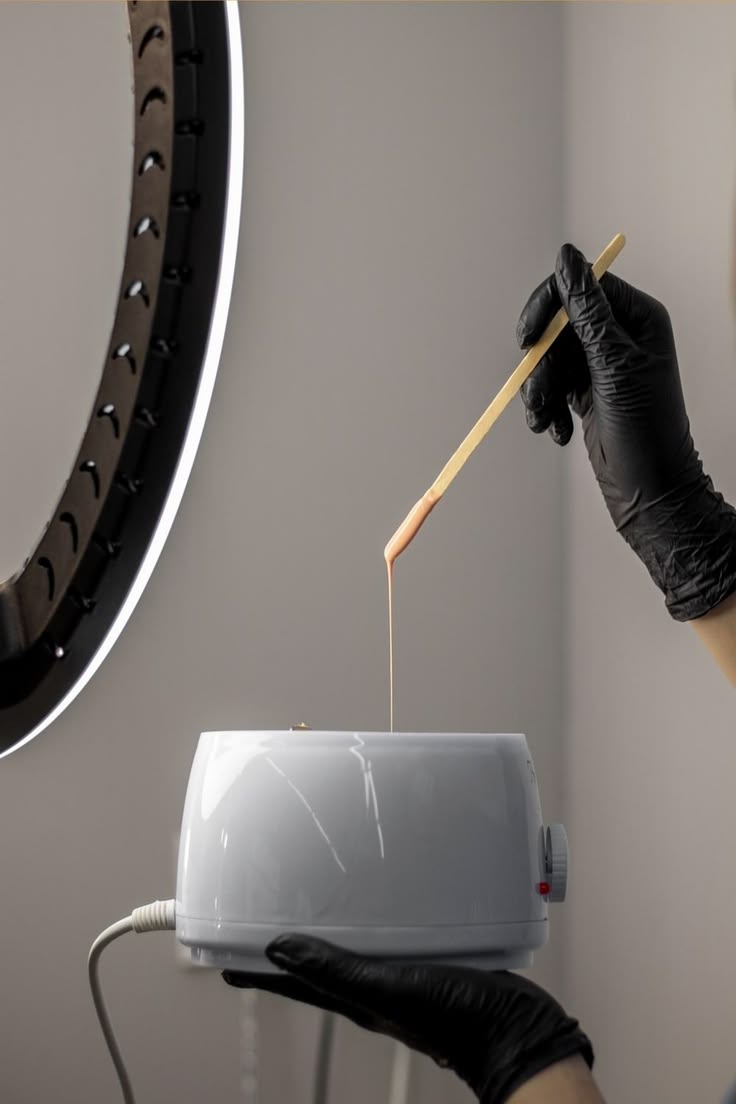 Mains gantées prélevant de la cire tiède à la spatule dans un chauffe-cire, préparation d'une épilation en cabine