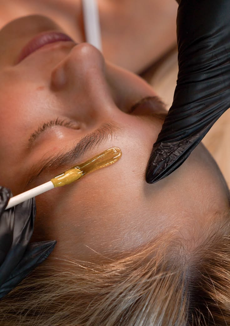 Esthéticienne gantée appliquant de la cire à la spatule sur le sourcil d'une cliente allongée en cabine