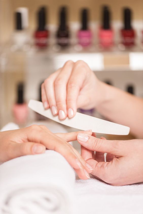 Gros plan sur les mains d'une élève qui lime les ongles d'une cliente pendant une manucure, flacons de vernis en arrière-plan